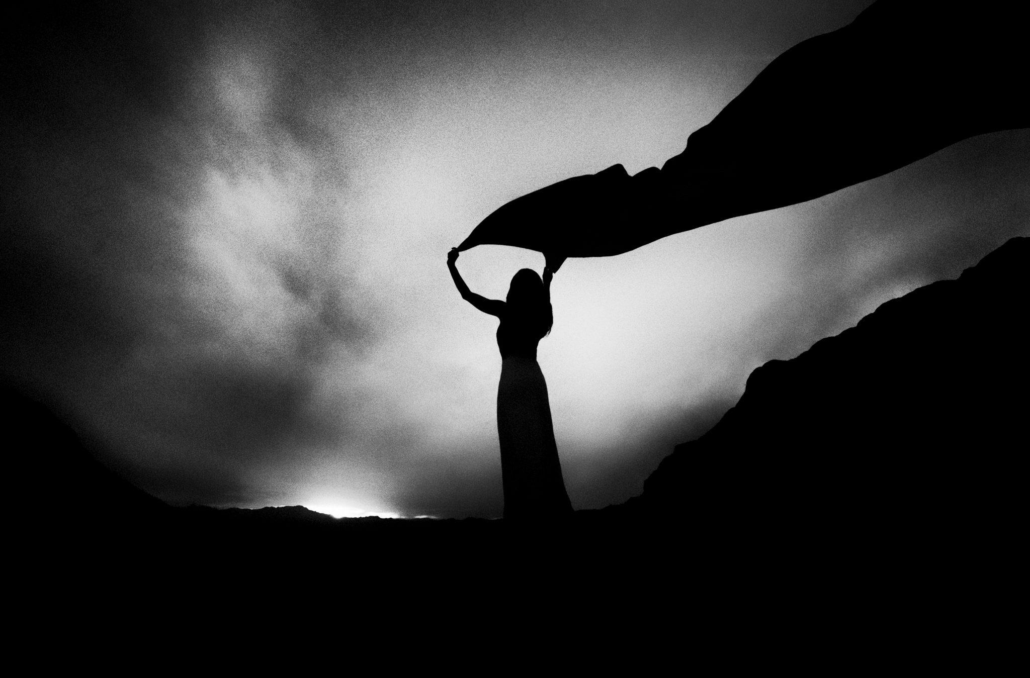 _ARS9594-Edit-Edit-Edit-4-Edit-2-Edit Black and white photo of a woman's sillouette holding a long scarf high over her head against a dawning sky.