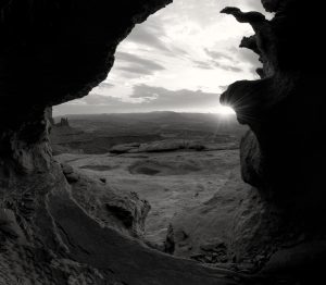 Black and whote phot of the interior of a cave looking out to the rising sun over the desert.