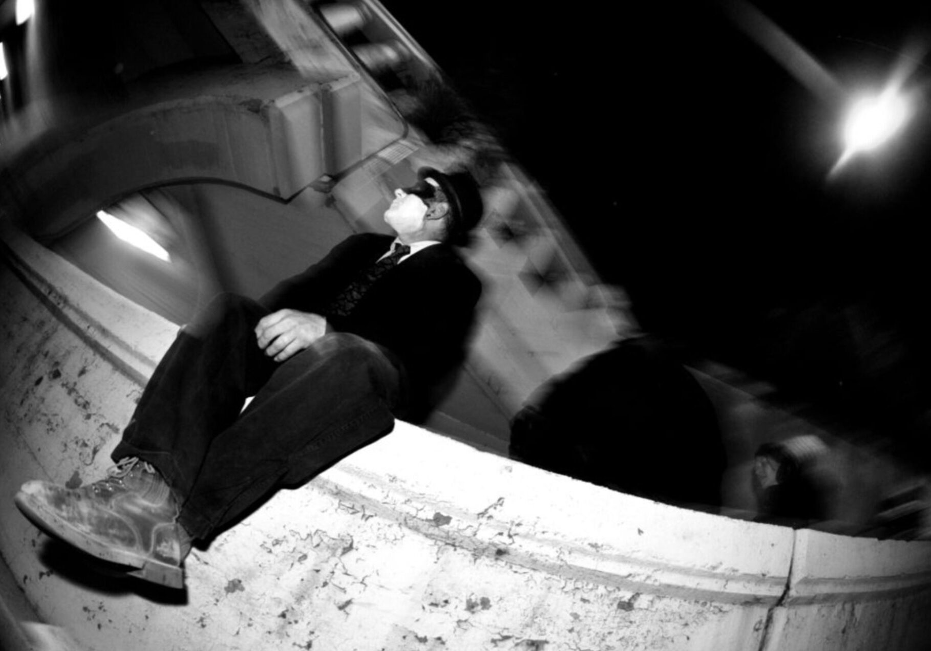 Screenshot 2023-04-15 at 4.36.51 PM Black and white photo of man with a white painted face sitting alone on a stone wall.